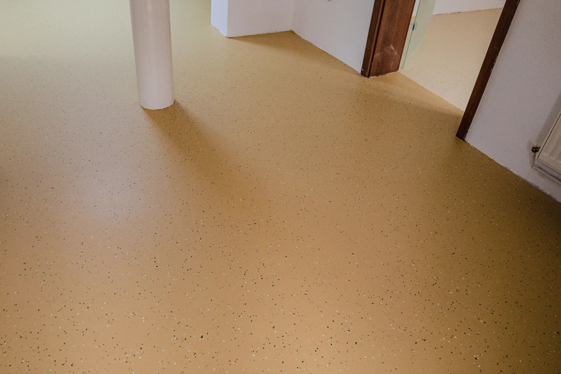 A room with a smooth, light brown floor with small speckles, white walls, a white pillar, and doorways leading to other rooms.
