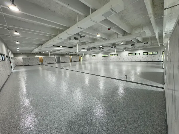 Large empty indoor space with a shiny, speckled gray floor, white walls, high ceiling with exposed beams, LED lights, and several small rectangular windows along one side.