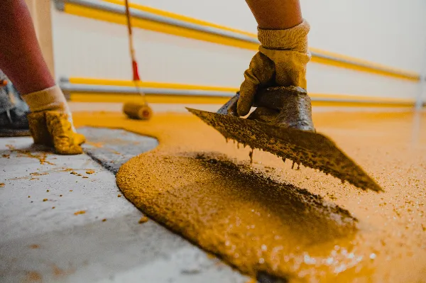 A close-up of a person’s gloved hands spreading yellow epoxy coating onto a concrete floor with a trowel while another person uses a paint roller in the background.