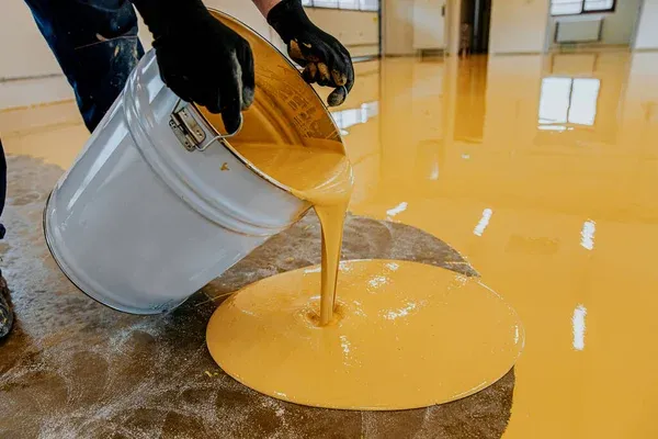 A person wearing black gloves pours yellow epoxy resin from a large silver bucket onto a floor, spreading the liquid to coat the surface in a brightly lit indoor space.