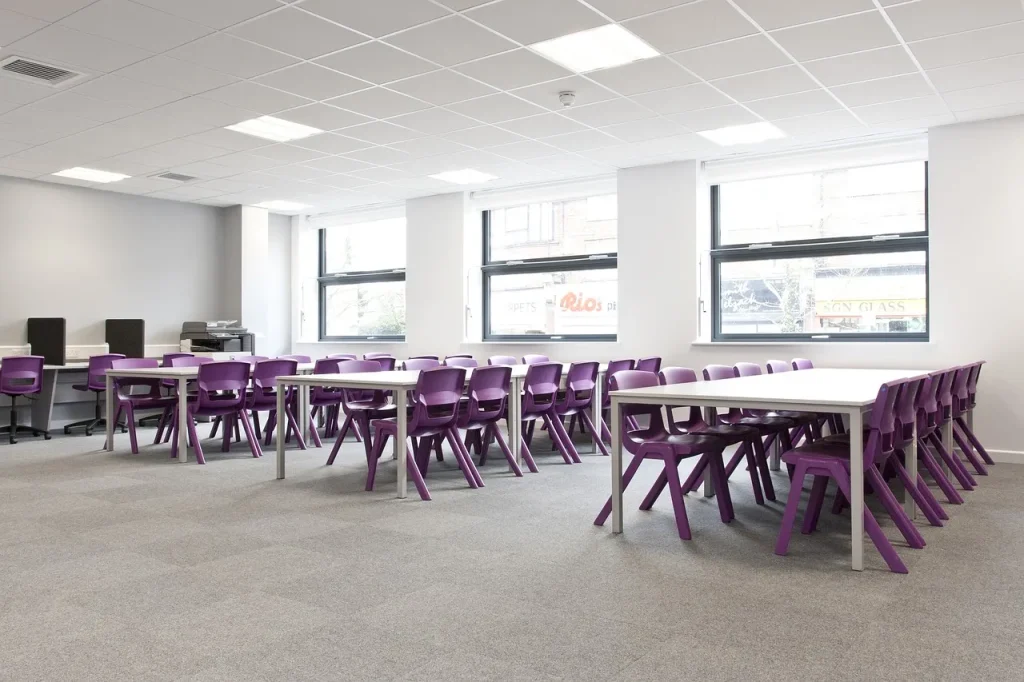 A modern classroom with large windows, white walls, and several tables surrounded by purple chairs. The room is empty, and computers are visible in the back corner. Natural light fills the bright, spacious area.