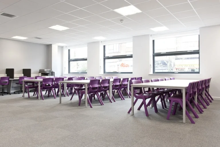 A modern classroom with rows of white tables and purple chairs, large windows letting in natural light, and a neutral gray carpet. There are computers and a printer in the back corner.