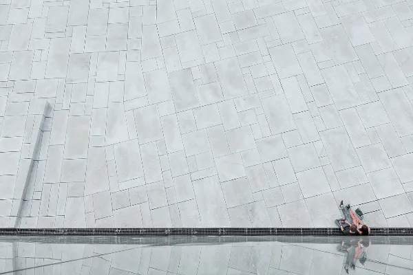 A person sits on a scooter at the edge of a large, light gray tiled plaza, with geometric patterns dominating most of the image and creating a minimalist, expansive feel.