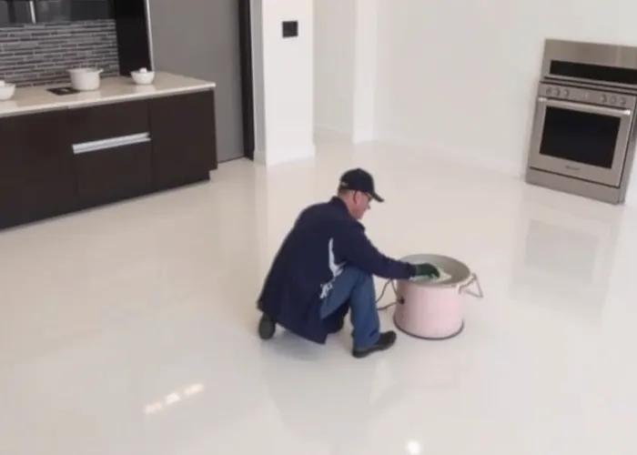 A person in a cap and jacket kneels on a shiny white floor, cleaning it with a round floor buffer in a modern, unfurnished kitchen with dark cabinets and a stainless steel oven.