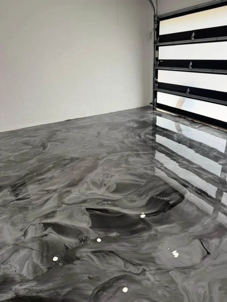 A garage with a shiny, swirl-patterned Miami Epoxy Resin floor in shades of gray and black. The garage door is partially open, and some small white objects are scattered on the smooth surface.
