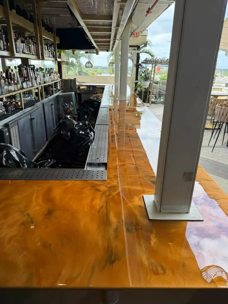 A shiny, marbled Miami Epoxy Resin amber-colored bar countertop reflects light indoors, with bottles and bar tools on one side and outdoor seating with palm trees visible through large windows in the background.