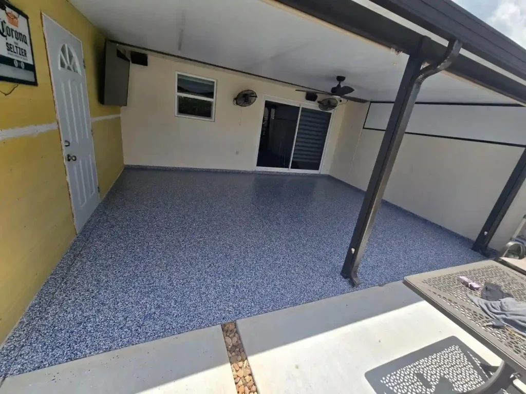 A covered outdoor patio with a speckled blue Miami Epoxy Resin floor, yellow and white walls, ceiling fans, and a door on the left. A table is partly visible in the foreground. The space appears clean and well-maintained.