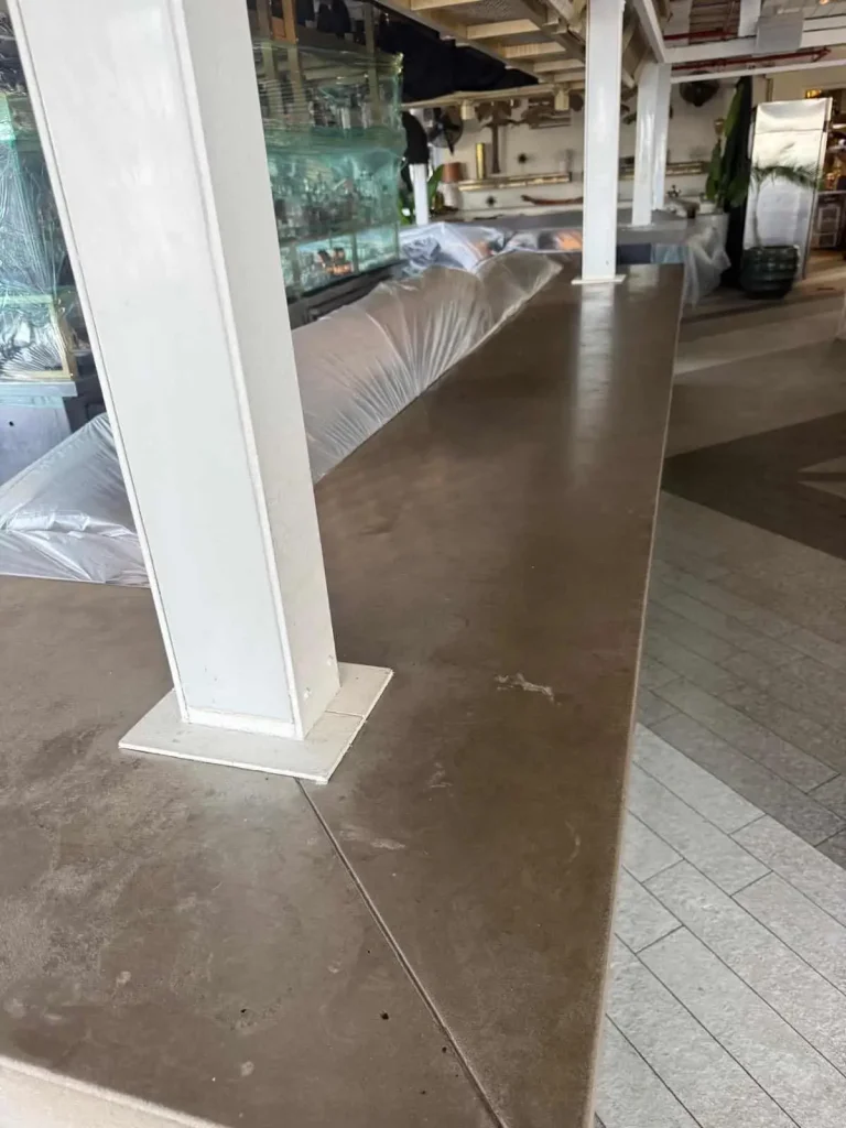 A long brown Miami Epoxy Resin countertop with a white support beam, next to a glass display filled with bottles. Plastic sheets cover objects in the background, and the floor has light gray tiles.
