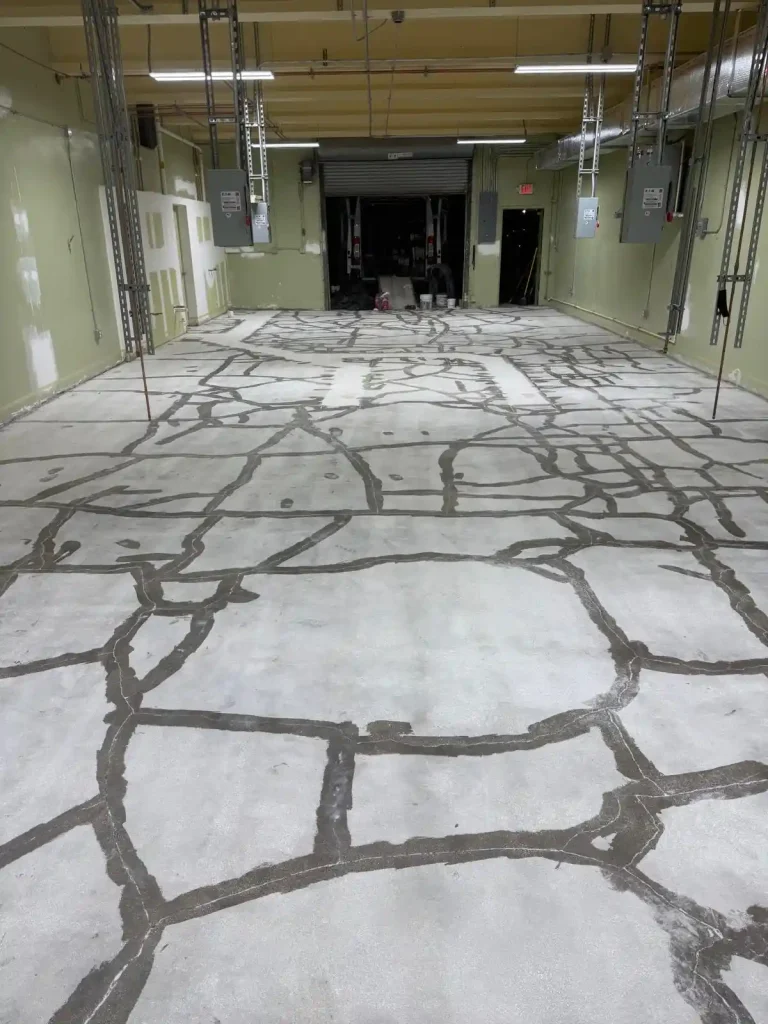 A large indoor room with a concrete floor covered in numerous dark, irregularly shaped cracks that have been patched or filled using Miami Epoxy Resin. The space features a high ceiling, exposed pipes, and a partially open garage door at the far end.