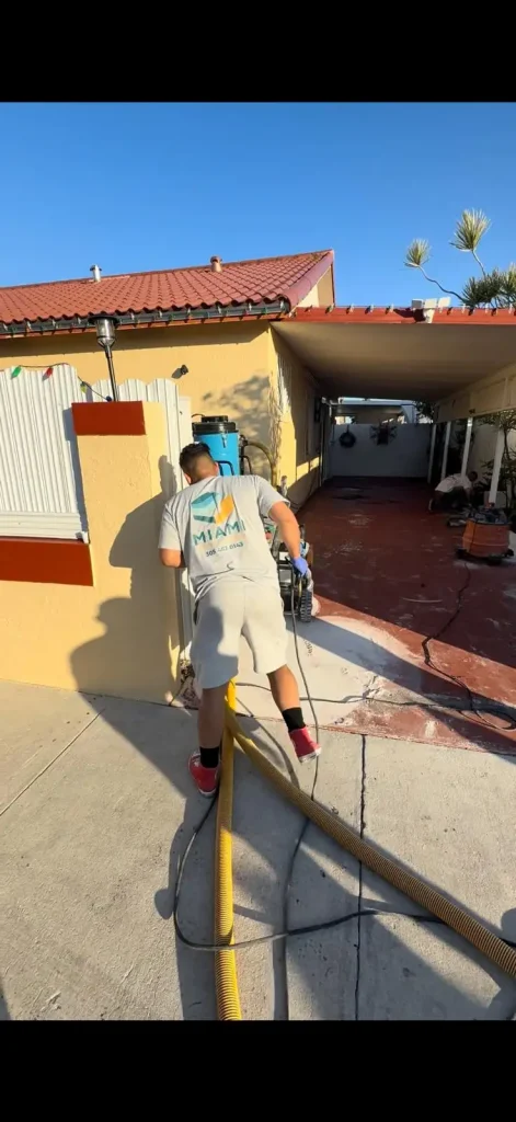 A person wearing a gray "Miami Epoxy Resin" t-shirt and shorts is working outside a house, handling a large yellow hose on a driveway near a carport on a sunny day.