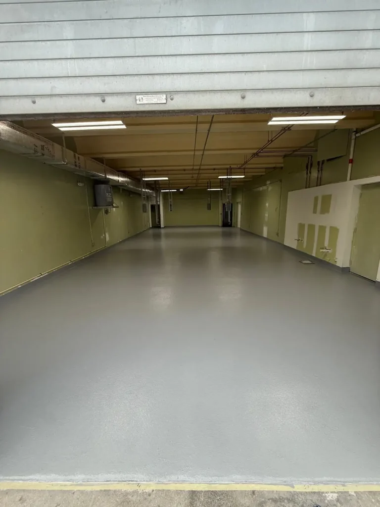 A wide, empty indoor garage with Miami Epoxy Resin gray floors, pale green walls, ceiling lights, and exposed pipes, viewed through a partially opened metal shutter door.