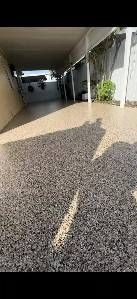 A covered outdoor patio with a speckled Miami Epoxy Resin floor, partially shaded. Plants line the right side near a white fence, and hanging planters are visible at the far end under the roof.