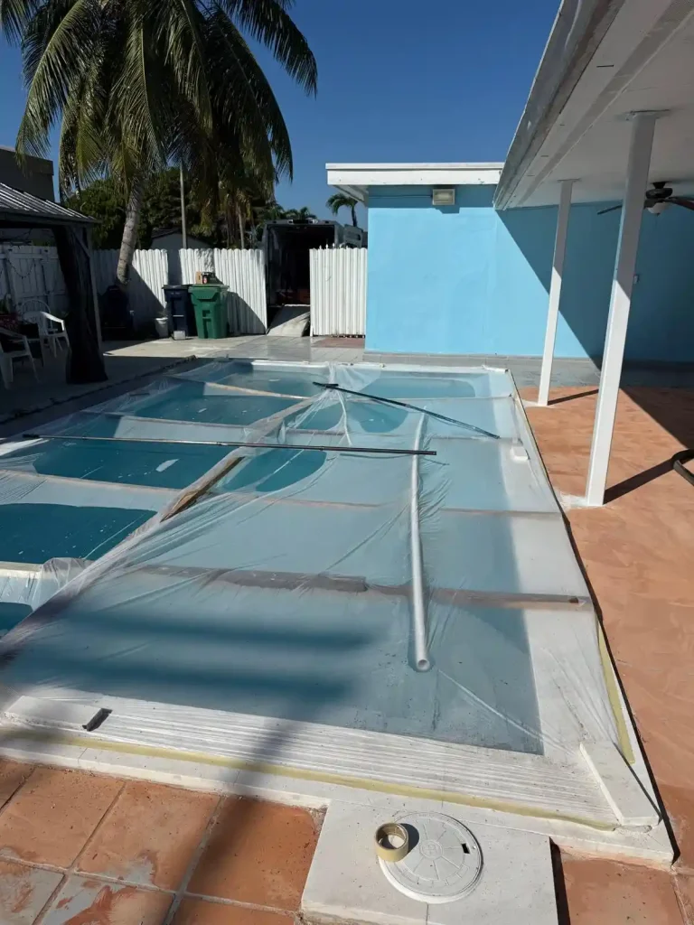 A backyard swimming pool is partially covered with clear plastic and metal poles. A palm tree, a blue shed, and some trash bins are visible in the background under a clear blue sky, evoking a Miami Epoxy Resin vibe.