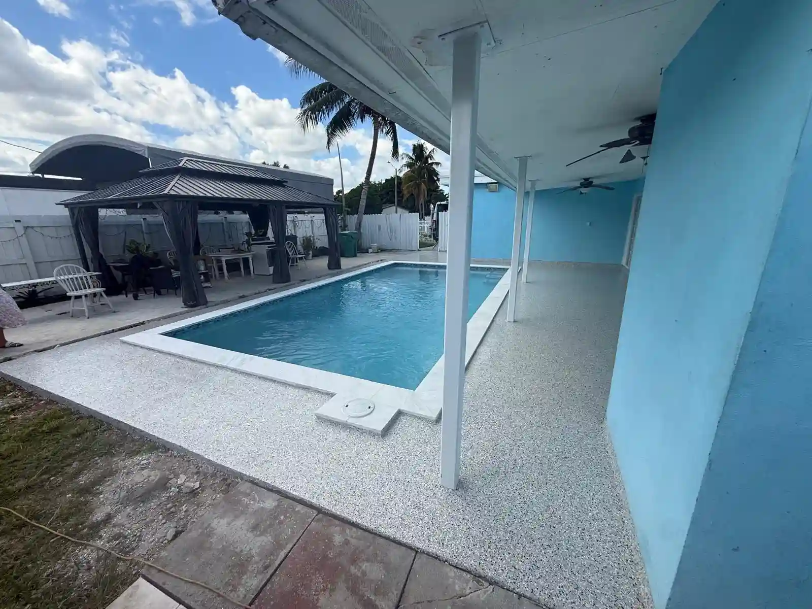 Rectangular swimming pool next to a light blue house, partially covered by a roof, with a gazebo, outdoor seating, palm trees, and Miami Epoxy Resin finishes for added style—all under a cloudy sky backdrop.