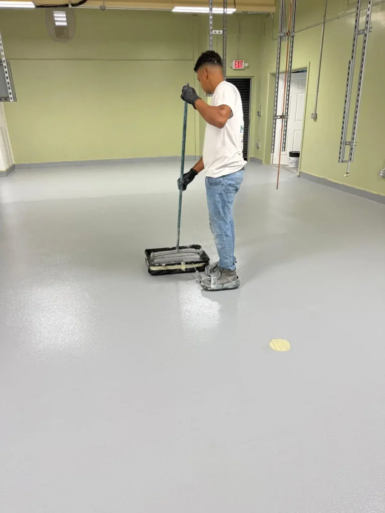 A person wearing gloves, jeans, and a white shirt is using a roller to paint or coat a large, light gray floor inside a spacious room with light green walls and exposed ceiling pipes.
