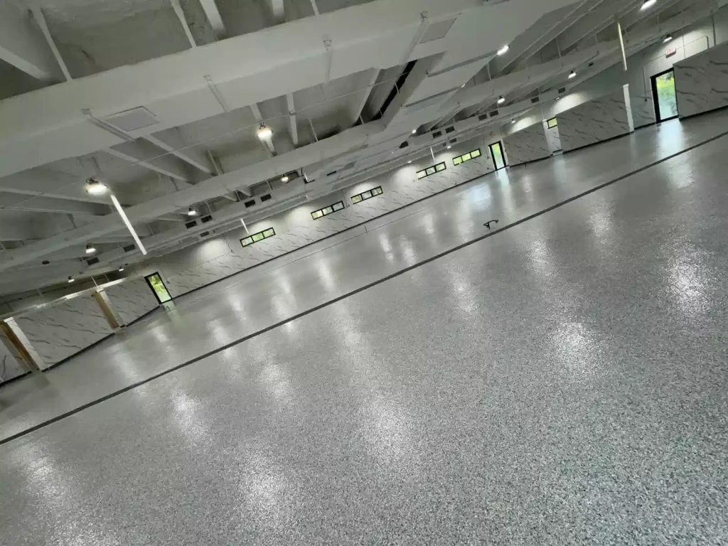 A spacious, empty room with a polished, speckled Miami Epoxy Resin floor, modern white ceiling, and walls featuring small windows and geometric designs. Ceiling lights are evenly spaced, with natural light streaming through several windows.