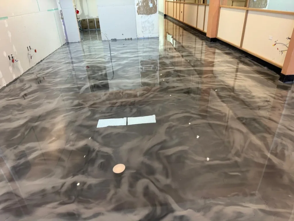 A room with a glossy, newly installed Miami Epoxy Resin floor featuring a swirling gray and black marble-like pattern; some construction materials and debris are visible on the floor.