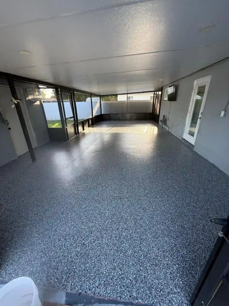 A covered patio with a speckled Miami Epoxy Resin floor, enclosed by dark-framed screens and doors. Sunlight enters from the left, and a door, window, and mounted TV are visible on the right wall.