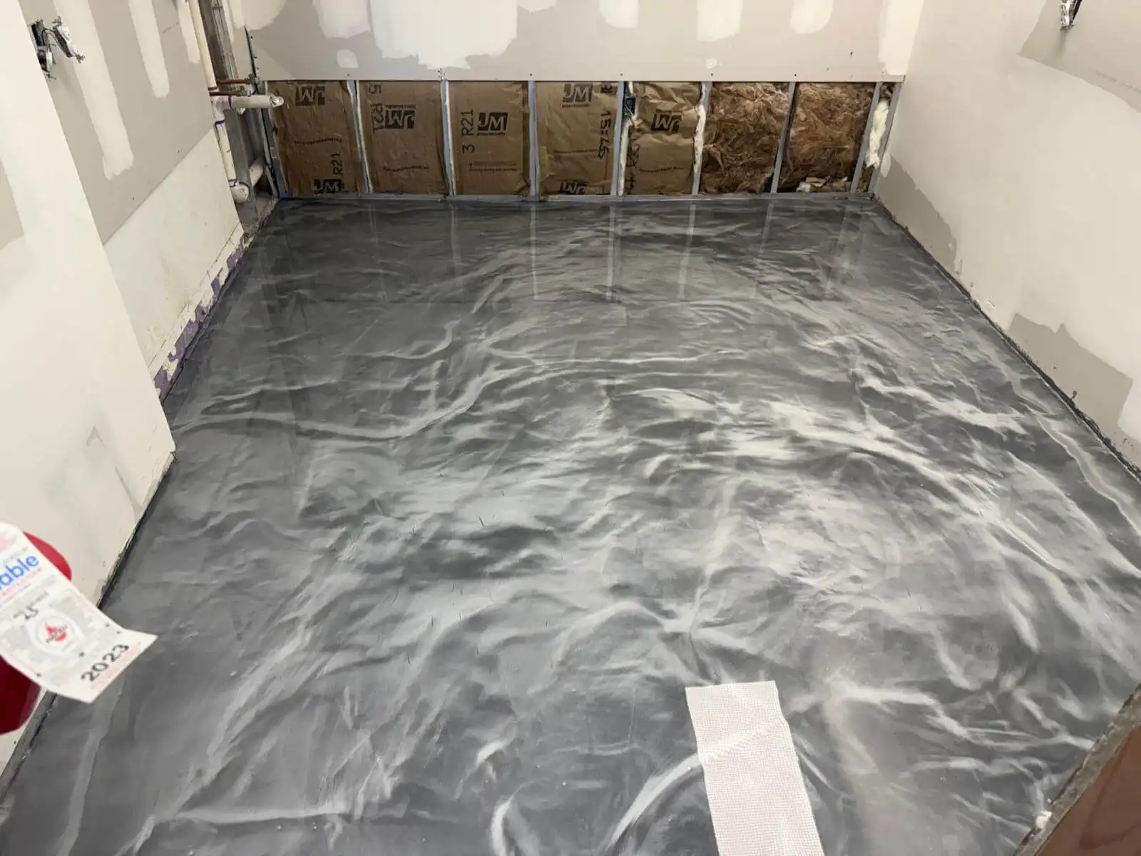 A room under renovation with unfinished drywall and exposed insulation, featuring a newly installed shiny gray Miami Epoxy Resin floor with elegant swirled patterns.