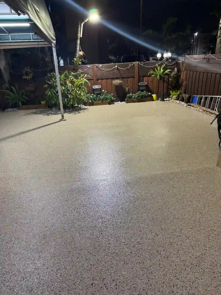 A clean, freshly resurfaced patio at night with Miami Epoxy Resin, surrounded by plants, a brown wooden fence, chairs, and outdoor lighting. Some tools and a ladder are visible by the fence.