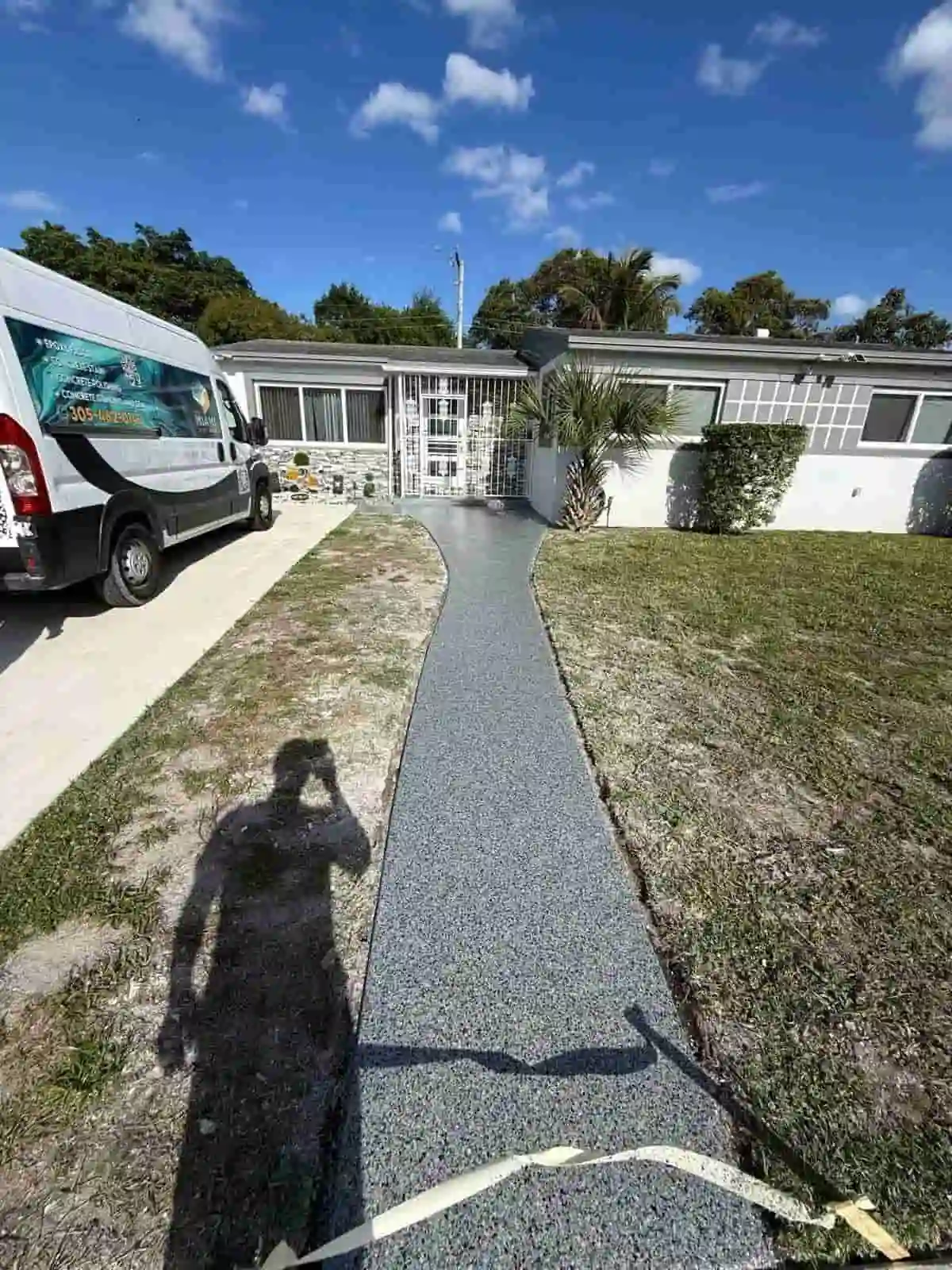 A narrow, curved gray pathway leads to a single-story house with white walls and large windows. A white van is parked in the driveway, and a person’s shadow is visible on the path. The yard has patches of grass.