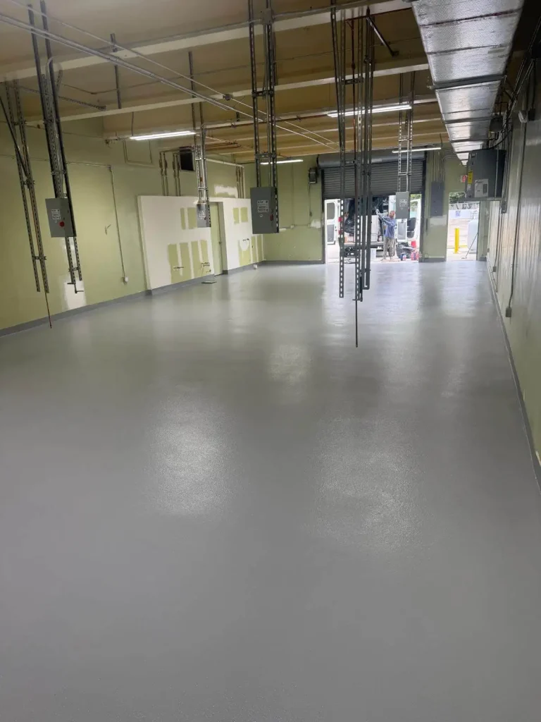 Empty industrial room with a smooth, light gray Miami Epoxy Resin floor, exposed pipes and electrical panels on the ceiling, pale green walls, and a large open doorway at the far end letting in daylight.