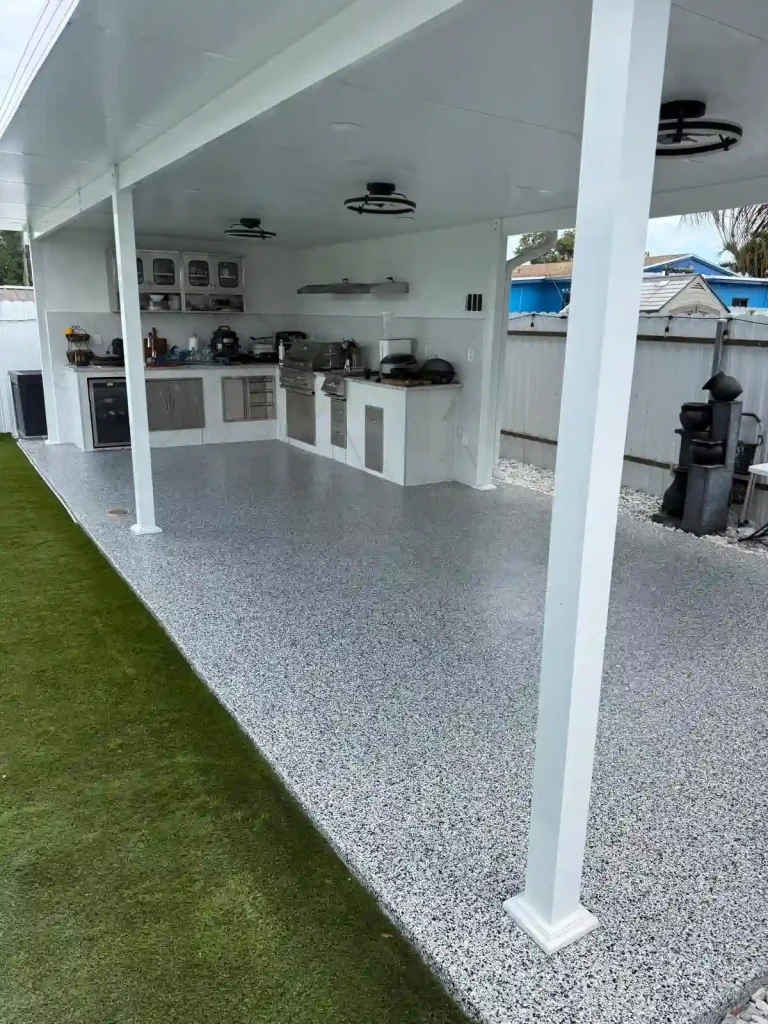 A modern outdoor kitchen with a Miami Epoxy Resin speckled gray floor, white ceiling, and exposed columns. The kitchen features stainless steel appliances, cabinets, and grill, with artificial grass elegantly bordering the area.