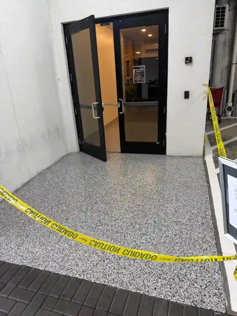 A building entrance with double glass doors is partially blocked by yellow caution tape, warning people to avoid the freshly finished Miami Epoxy Resin speckled floor surface in front of the doorway.
