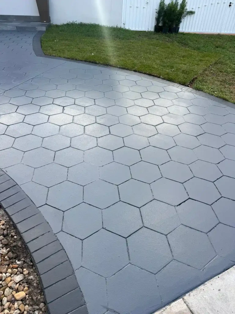 A walkway with large gray hexagonal pavers curves through a yard, bordered by dark rectangular bricks and green grass. Stones line one edge of the path.