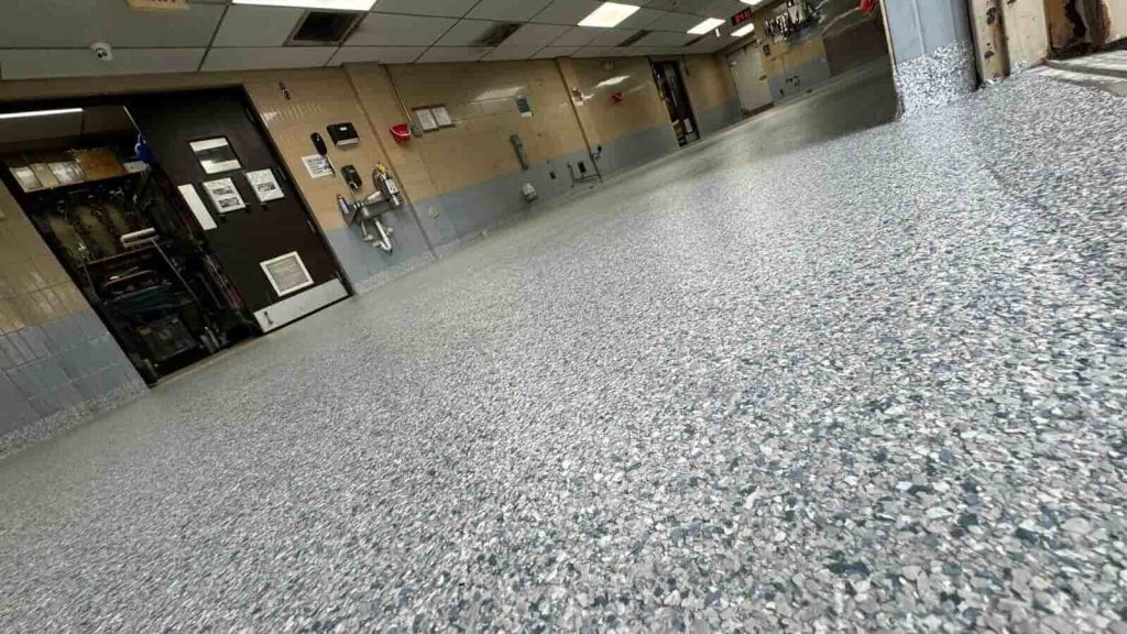 Low-angle view of a clean, speckled Miami Epoxy Resin floor in an industrial or commercial room with beige and blue tile walls, overhead fluorescent lights, and various equipment and doors along the walls.