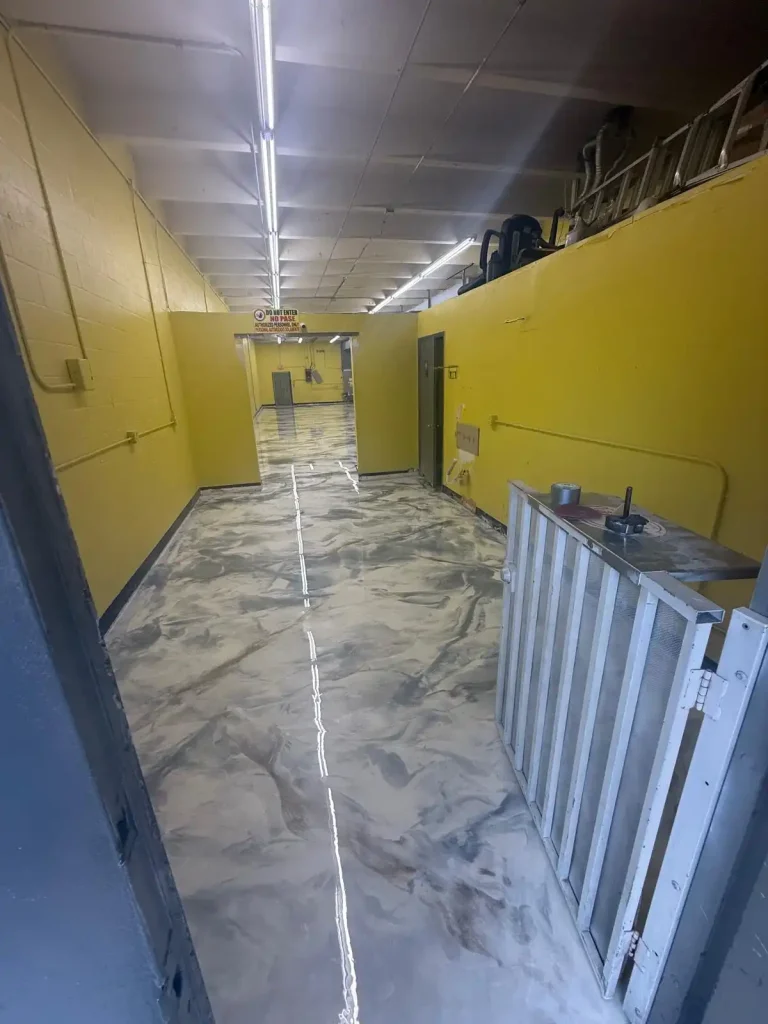 A hallway with bright yellow walls and overhead fluorescent lights features a shiny, marbled gray and white Miami Epoxy Resin floor. There is a wooden barrier and some equipment on the right side near the entrance.